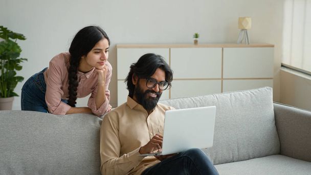 Arabian Indian couple on couch use laptop computer shopping online together pay bills order food at home choose buying internet goods give high five gesture happy man and woman approve teamwork unity - Photo, Image