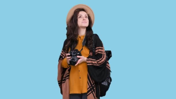 Elegante jovem mulher de chapéu com mochila vai fazer foto por câmera, olhando em torno de fundo isolado azul. Viagem, turismo, conceito de aventuras - Filmagem, Vídeo