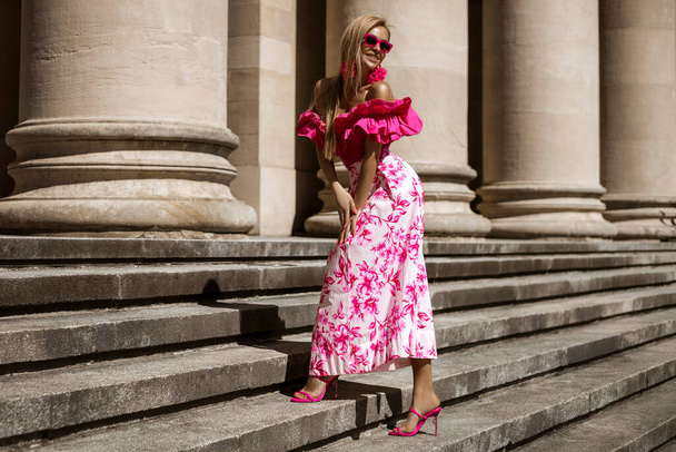 Elegantní, stylová žena v růžových květinových šatech a sexy vysokých podpatcích se smyslně prochází ve městě za slunečného letního dne. Venkovní natáčení. - Fotografie, Obrázek