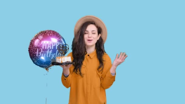 Smiling young woman posing with cake and balloon while celebrating her birthday on blue isolated background. Party concept - Footage, Video