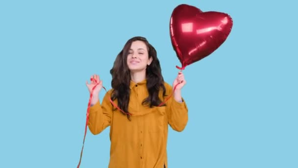 Smiling young woman posing with heart shape air balloon and dancing on blue isolated background. Love concept. Valentine Day - Footage, Video
