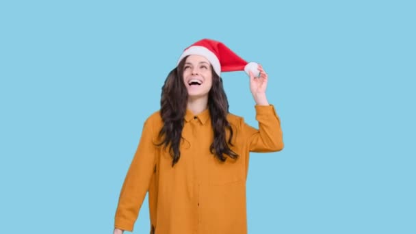 Счастливая молодая женщина в шляпе Санты танцует во время празднования Рождества на голубом изолированном фоне. Праздник, праздничная концепция - Кадры, видео