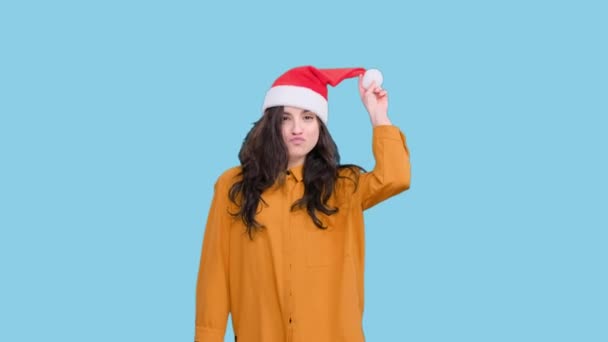 Glimlachende jonge vrouw in Santa hoed grimmig tijdens het vieren van Kerstmis op blauwe geïsoleerde achtergrond. Feestdag, vakantie concept - Video