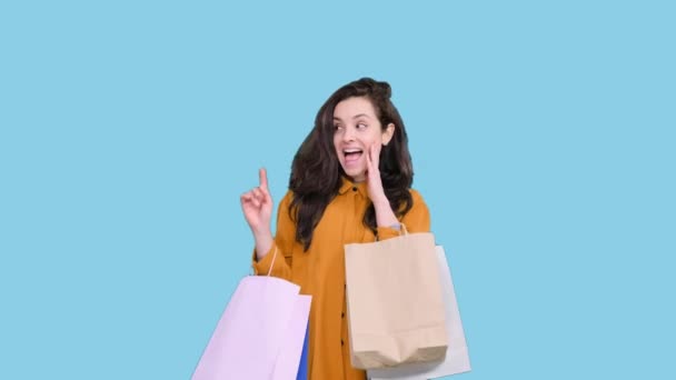 Alışveriş torbaları tutan mutlu genç bir kadın mavi izole arka plandaki boşluğu parmağıyla işaret ediyor. Kara Cuma, satış konsepti - Video, Çekim