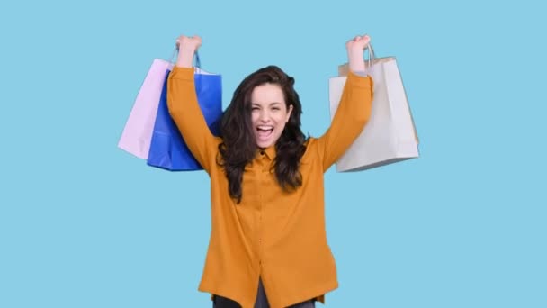 Eine fröhliche junge Frau hält bunte Einkaufspakete in der Hand, während sie sie auf blauem Hintergrund hochhält. Black Friday, Verkaufskonzept - Filmmaterial, Video