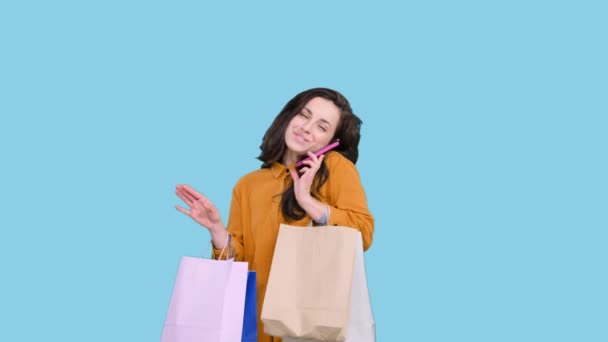 Улыбающаяся женщина с сумками, стоящая во время разговора по мобильному телефону на синем изолированном фоне. Черная пятница, концепция продажи - Кадры, видео