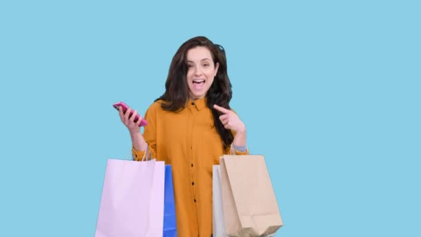 Jeune femme souriante posant avec des sacs à provisions tout en utilisant un smartphone, pointant l'écran sur fond bleu isolé. Black Friday, concept de vente - Séquence, vidéo