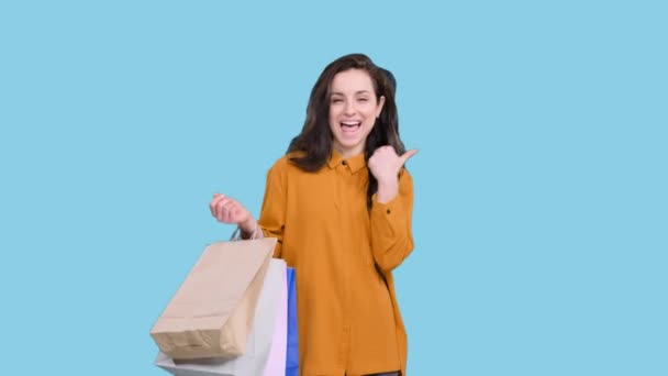 Улыбающаяся красивая женщина с бумажными пакетами, указывающими пальцем на пространство для копирования на синем изолированном фоне. Черная пятница, концепция продажи - Кадры, видео