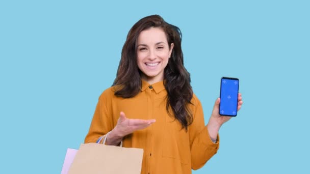 Vrolijke vrouw vrouw met kleurrijke boodschappentassen terwijl het houden van slimme telefoon met kopieerruimte op het scherm op blauwe geïsoleerde achtergrond. Black Friday, verkoopconcept - Video