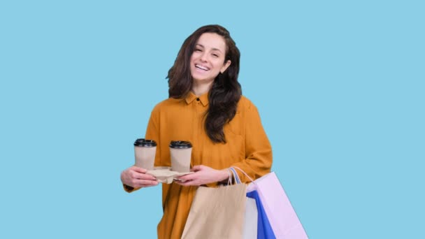 Jovencita feliz disfrutando de las compras, sosteniendo bolsas y dos tazas de café sobre fondo azul aislado. Viernes Negro, concepto de venta - Imágenes, Vídeo