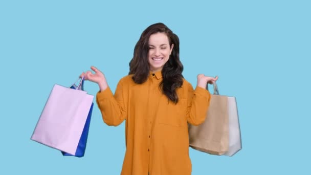 Uśmiechnięta ładna kobieta niosąca kolorowe torby na zakupy, ciesząc się zakupami na niebieskim odosobnionym tle. Black Friday, koncepcja sprzedaży - Materiał filmowy, wideo