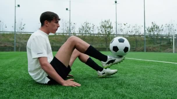 Підлітковий футболіст робить футбольні фрістайли сидячи в полі. - Кадри, відео