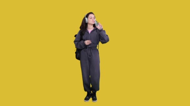 Smiling woman listening the music on the headphones while posing with backpack and drinking water on yellow isolated. Full length - Footage, Video
