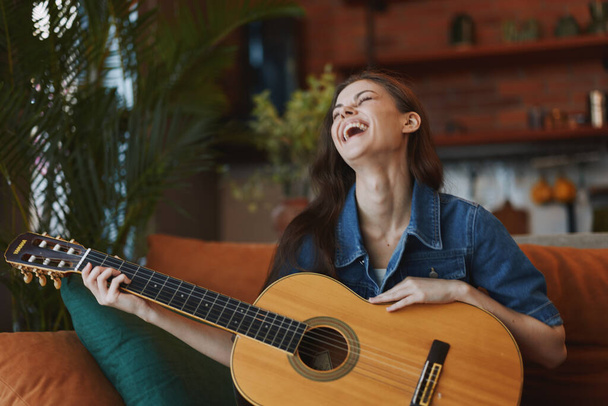 Glückliche Frau spielt Akustikgitarre und lacht, während sie auf einer Couch im gemütlichen Wohnzimmer sitzt - Foto, Bild