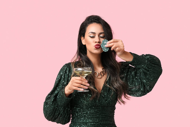 Bella giovane donna afro-americana con un bicchiere di champagne baciare chip di poker su sfondo rosa - Foto, immagini