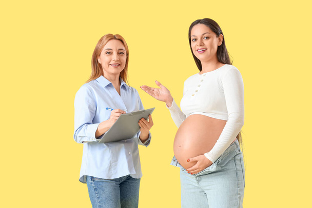 成熟したドゥーラ 黄色い背景の妊婦とクリップボードで書く - 写真・画像
