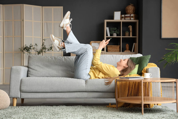 Mladá žena pomocí mobilního telefonu na šedé pohovce v obývacím pokoji - Fotografie, Obrázek