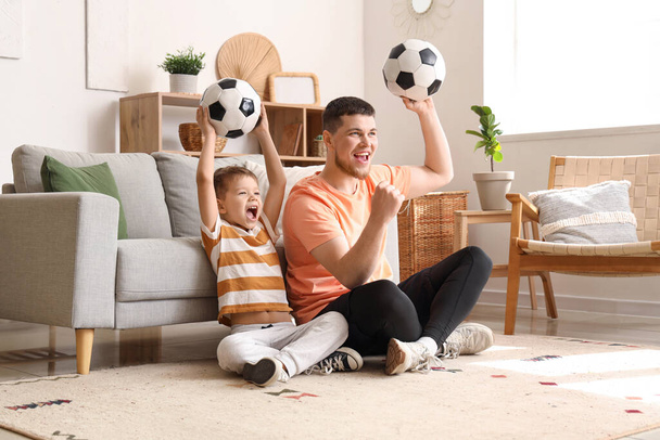 Young man and his little son cheering for football team with soccer balls at home - Photo, Image