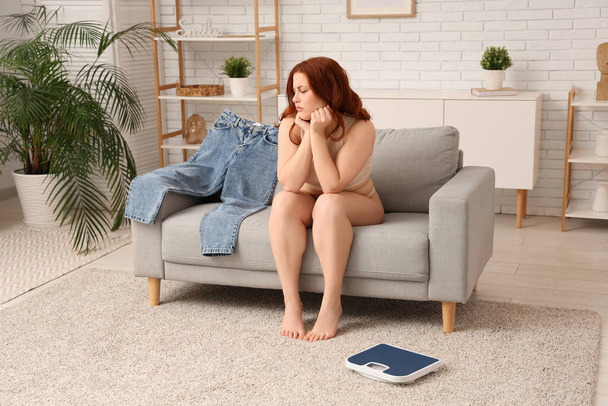 Järkyttynyt nuori nainen, jolla on tiukat farkut, istuu sohvalla kotona. Painonnousu käsite - Valokuva, kuva