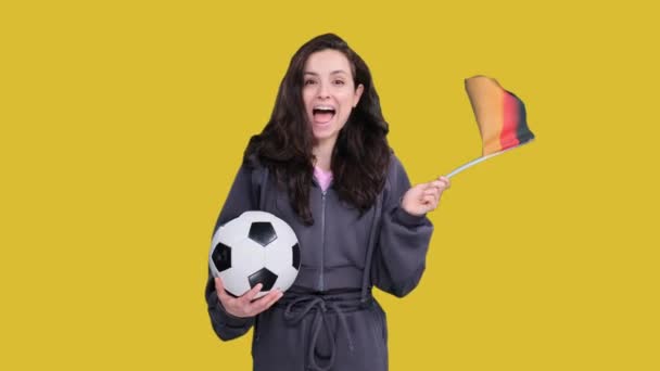 Happy young female fan posing with German flag while holding a soccer ball, celebrating team victory on yellow isolated - Footage, Video