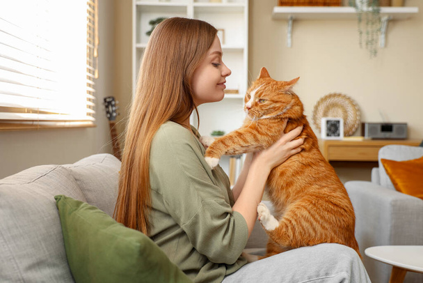 Mladá žena s roztomilou červenou kočkou sedí na pohovce doma - Fotografie, Obrázek