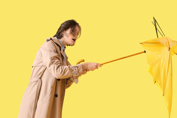 Mujer joven con paraguas roto y fuerte viento sobre fondo amarillo - Foto, Imagen