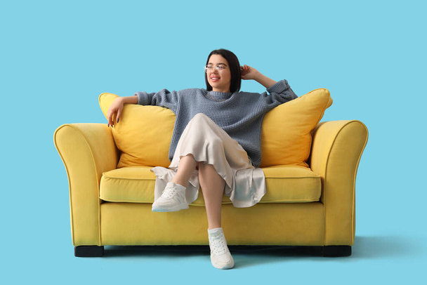 Young woman sitting on yellow sofa against blue background - Photo, Image