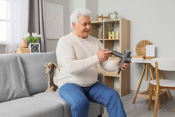 Starší muž s perkusní masážní sedící na pohovce doma - Fotografie, Obrázek