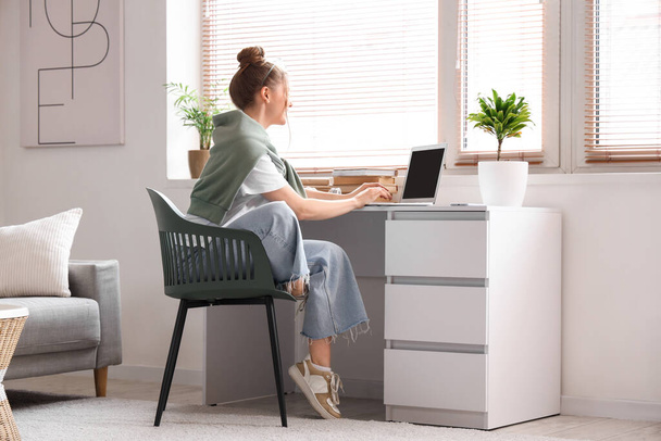Genç bir kadın iş yerinde oturuyor ve oturma odasında dizüstü bilgisayarla çalışıyor. - Fotoğraf, Görsel