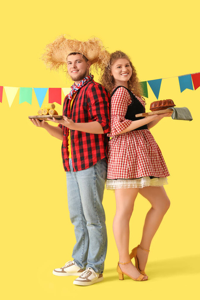 Feliz pareja joven con banderas, maíz y pastel sobre fondo amarillo. Celebración de Festa Junina - Foto, imagen