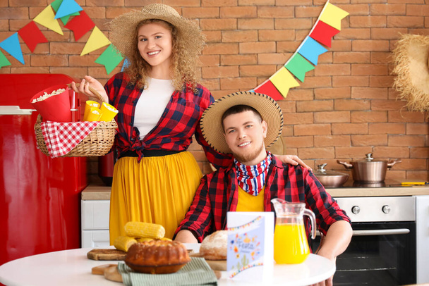 Feliz pareja joven sentada a la mesa con maíz, tarta y palomitas de maíz en la cocina. Celebración de Festa Junina - Foto, Imagen