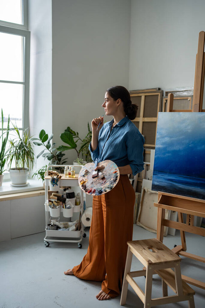 Zamyšlená umělkyně hledající inspiraci. Pensive toužebná žena drží štětce, paleta stojí vedle stojanu s kresbou, dívá se z okna, přemýšlet o dalším kroku v umělecké tvorby - Fotografie, Obrázek