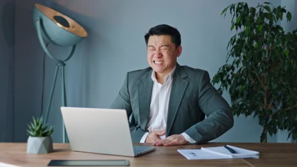 Jóképű kínai férfi hasi fájdalom a szünetben a munkahelyen hordozható laptoppal és fényes lámpával a háttérben. Frusztrált fiatal üzletember megérinti gyomor nyugodt irodai környezetben. - Felvétel, videó