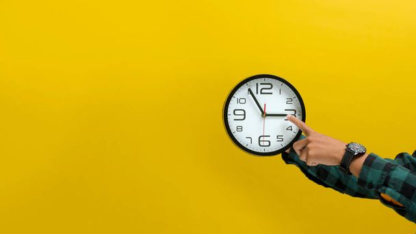 Mano sosteniendo un reloj de pared sobre un fondo amarillo brillante. Ideal para ilustrar la gestión del tiempo, recordatorios o el concepto de cuenta regresiva. - Foto, Imagen