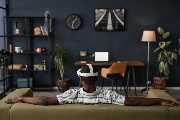 Вид сзади на неузнаваемого чернокожего человека в гарнитуре, сидящего расслабленно на диване в гостиной, пространство для копирования - Фото, изображение