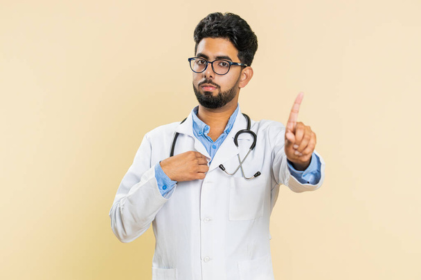 I am dont want it, not me. Confused Indian young doctor cardiologist man pointing fingers ask say who why me no thanks i do not need it rejection refusal stop sign. Apothecary guy on beige background - Photo, Image