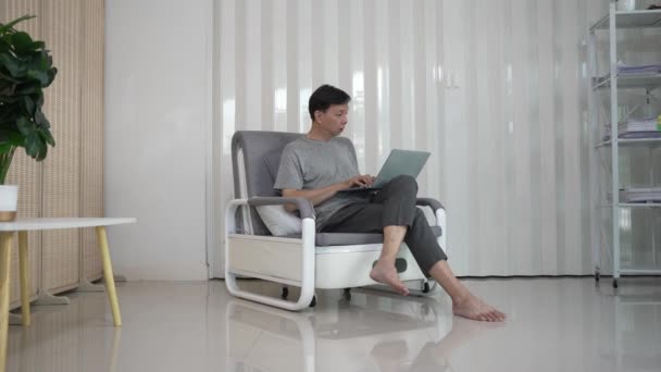 Oudere Aziatische man die zich vermaakt met zijn laptop computer, social media checkt en nieuws leest of online winkelt terwijl hij thuis op de bank zit in de woonkamer. - Video