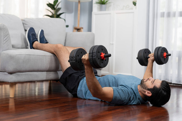 Athletic body and active sporty man using furniture for effective targeting muscle gain with weight lifting dumbbell exercise at gaiety home as concept of healthy fit body home workout lifestyle. - Photo, Image