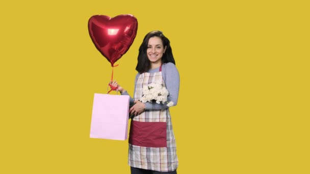 Elinde beyaz karanfiller, hediye çantası ve sarı izole edilmiş arka planda kırmızı kalp şeklinde balon tutan gülümseyen kadın çiçekçi. Küçük işletme kavramı - Video, Çekim