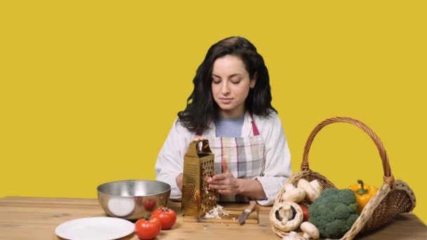 Pretty woman in apron sitting at the table while grating mushrooms and cutting her finger on yellow isolated background. Cooking, culinary, healthy lifestyle concept - Footage, Video