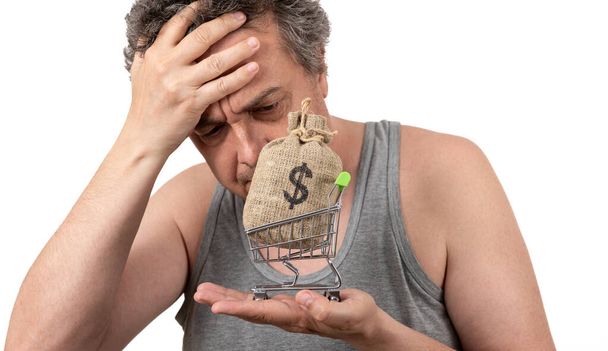 Un uomo rasato dai capelli grigi in una maglietta senza maniche tiene un carrello della spesa in miniatura da un supermercato e un sacchetto di denaro con un simbolo del dollaro USA - Foto, immagini