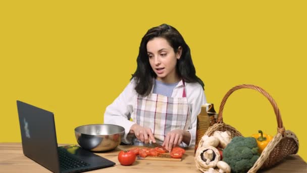 Pretty young woman using laptop while cooking, slicing tomato and talking online on yellow isolated background. Cooking, culinary, healthy lifestyle concept - Footage, Video