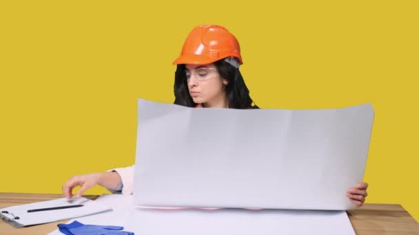 Mujer joven con casco protector trabajando con dibujo, sentada en el escritorio y tomando notas sobre fondo amarillo aislado. Industria, concepto de ingeniería - Metraje, vídeo