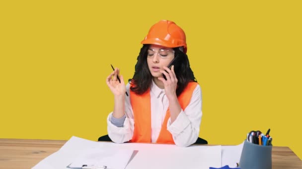 Mladá žena inženýrka v ochranné přilbě mluví na smartphonu, zatímco si dělá poznámky na žlutém izolovaném pozadí. Průmysl, inženýrský koncept - Záběry, video