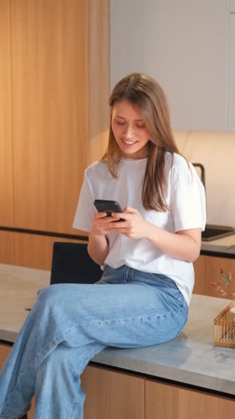 Вертикальне відео жінки, сидячи на кухні і використовуючи свій смартфон і посміхаючись. - Кадри, відео