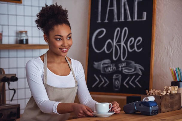 Beautiful Afro American barista in apron is holding a cup and smiling while standing at bar counter - Photo, Image