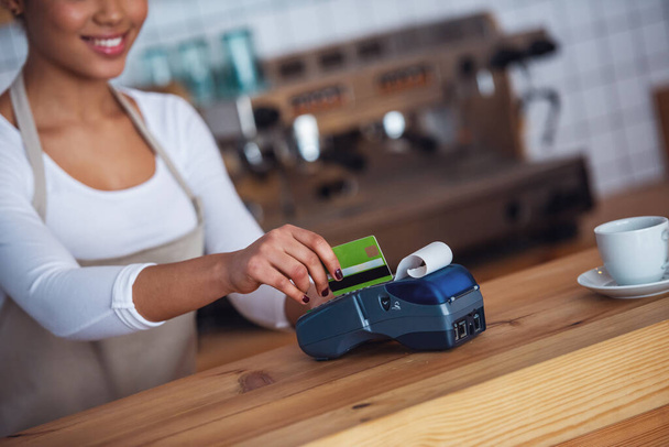 Imagem recortada da bela barista afro-americana em avental usando um terminal de pagamento e sorrindo enquanto estava de pé no balcão do bar
 - Foto, Imagem