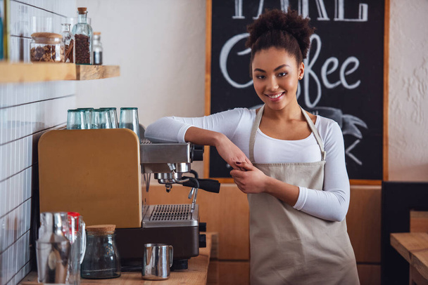 Bonito barista afro-americano em avental está olhando para a câmera e sorrindo enquanto se inclina na máquina de café
 - Foto, Imagem