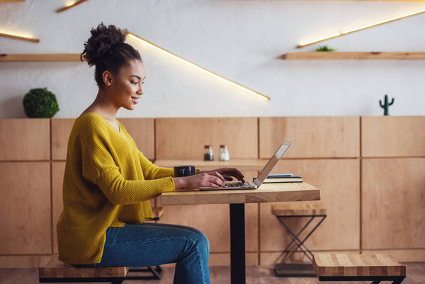 A szép Afro-amerikai lány alkalmi ruhák, mosolyogva, miközben dolgozik a laptop a kávézóban oldalnézete - Fotó, kép
