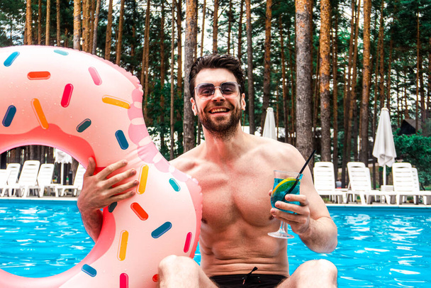 Усміхнений молодий бородатий чоловік в сонцезахисних окулярах з надувним кільцем у басейні, тримає коктейль на відкритому повітрі. Літо, свята, концепція способу життя - Фото, зображення